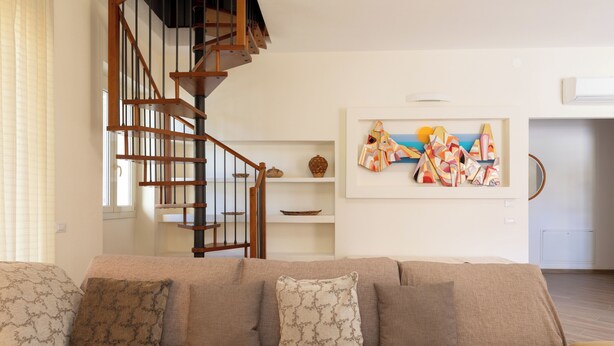 A living room with a beige sofa, a wooden staircase, and a wall-mounted abstract art piece. - Casa Marina con Patio by Wonderful Italy (Olbia)