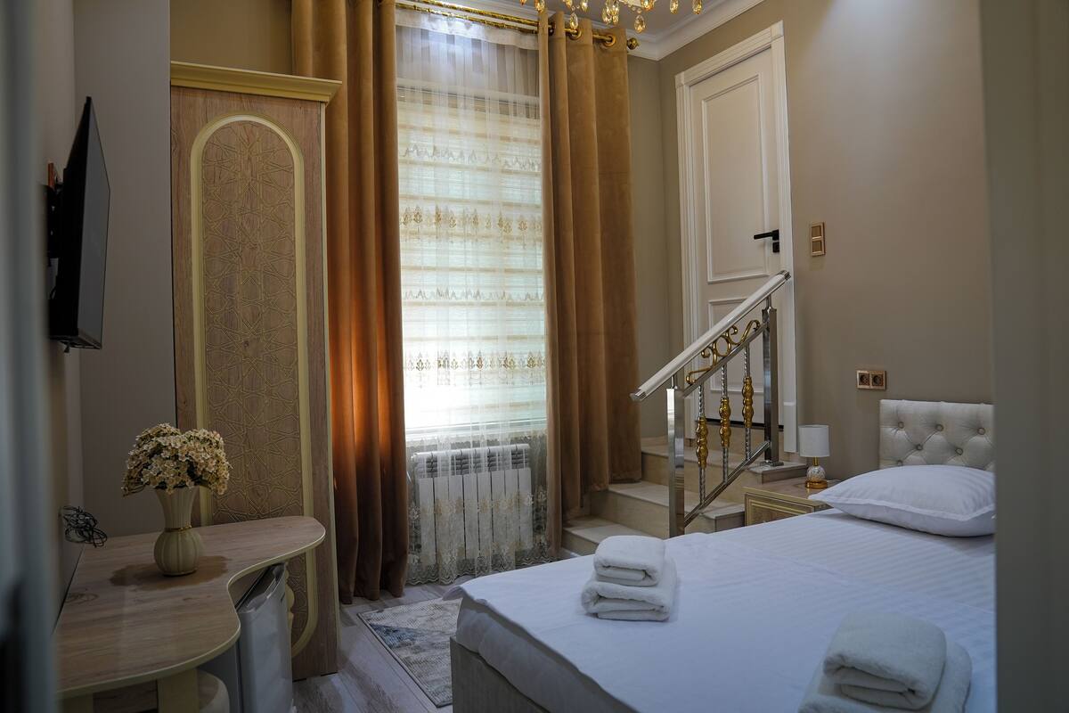 A hotel room with a bed, bedside table, lamp, and a view of the outside through a window with curtains.