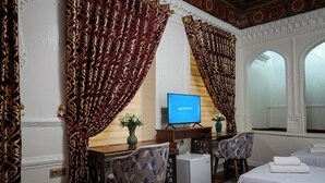 Habitación de hotel con dos camas, un escritorio con televisor, cortinas estampadas y un techo decorativo.