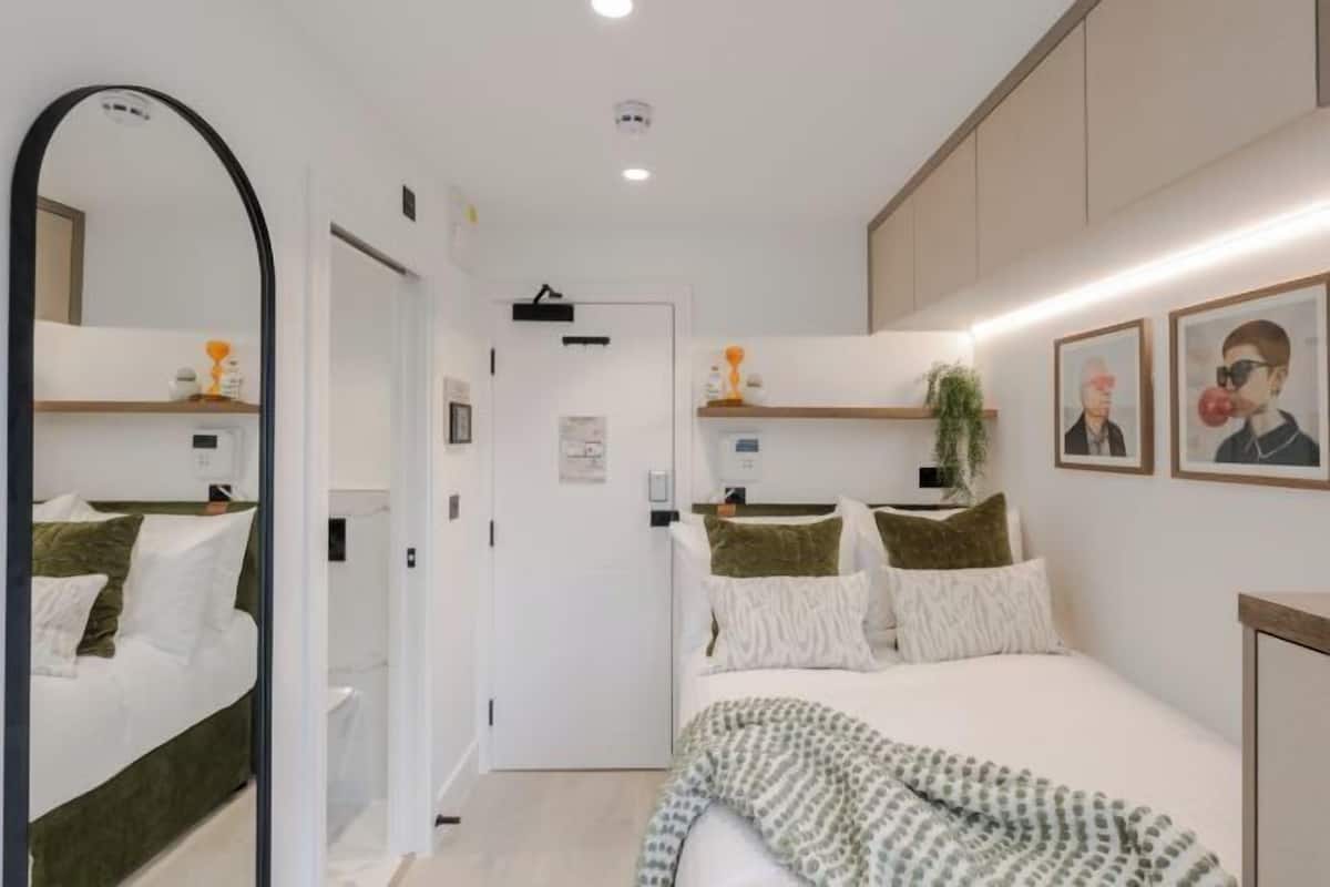 Una habitación de hotel moderna con una cama grande, un espejo, una mesita de noche con lámpara y un estante en la pared con objetos decorativos.