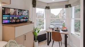 A modern room with a large TV on a cabinet, a desk with a chair, and large windows with curtains. - Kensington Standard Room (London)