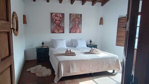 Une chambre avec une porte en bois, un lit recouvert de draps blancs, deux tableaux accrochés au mur et un petit tapis sur le sol.