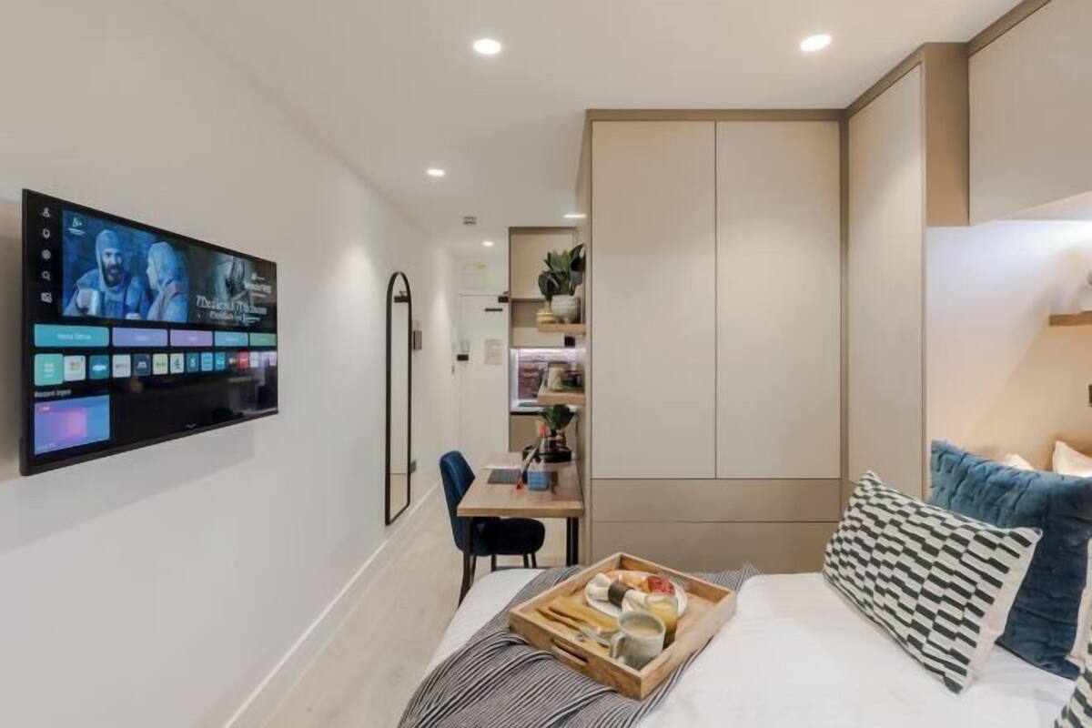 A modern hotel room with a flat-screen TV, a bed with a tray of breakfast items, a desk, and a wardrobe.