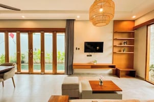 A modern living room with a flat-screen TV, wooden shelves, and a large window overlooking a pool.