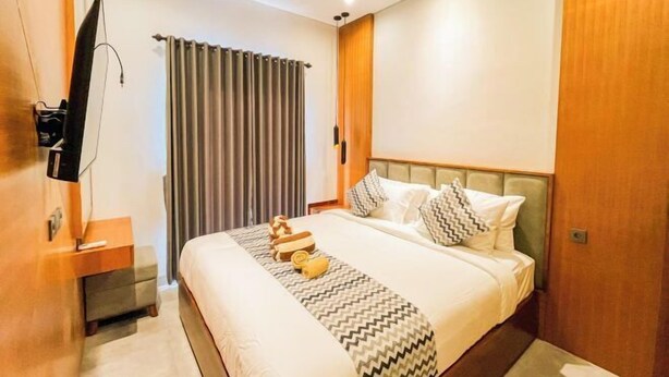 A modern hotel room with a large bed, a flat-screen TV mounted on the wall, a wooden nightstand, and a window with curtains. - KANGIN House Sanur (Bali)