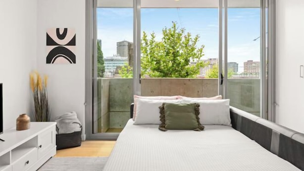Un dormitorio moderno con una cama grande, una puerta corrediza de vidrio a un balcón y vista a la ciudad.