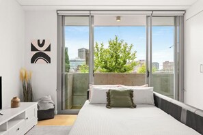 A modern bedroom with a large bed, a sliding glass door to a balcony, and a city view. - Central stylish 1 bedroom with balcony and air-con (Surry Hills)