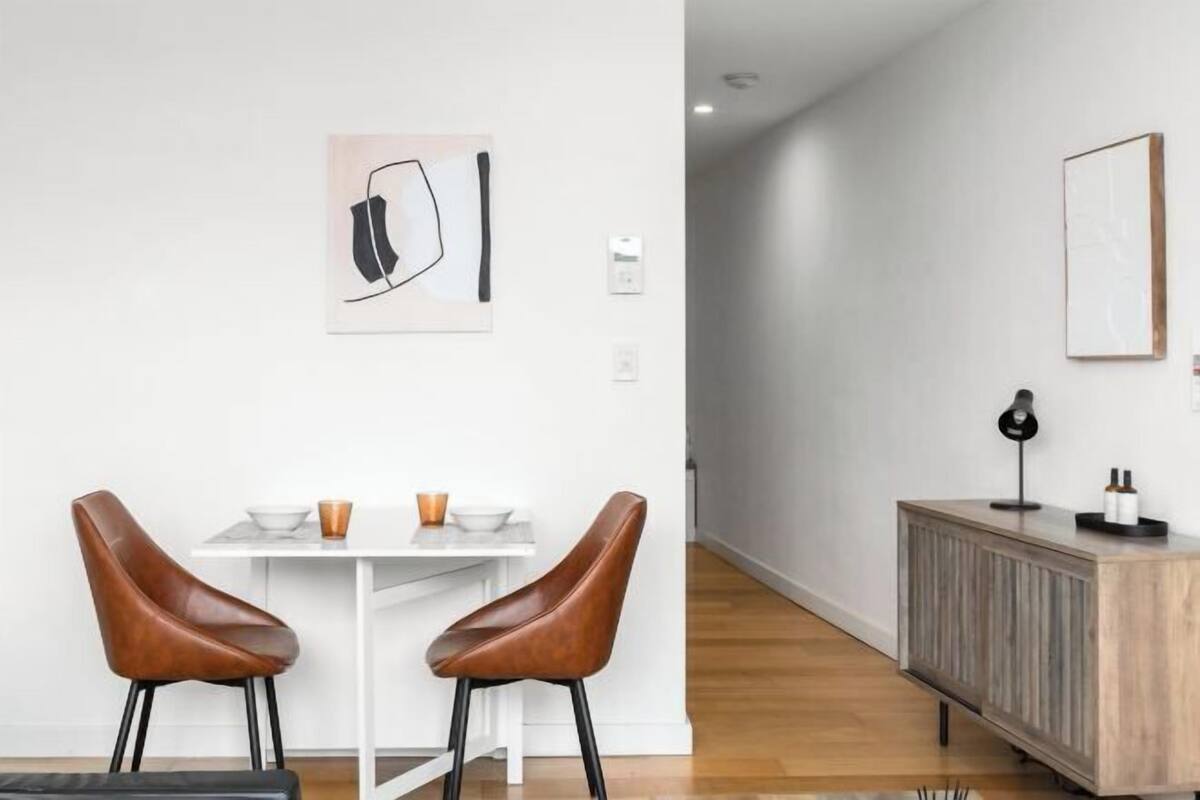 Un comedor moderno con dos sillones de cuero, una mesa blanca y una decoración minimalista que incluye un cuadro enmarcado y un aparador con una lámpara.