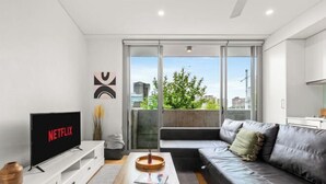 Una sala de estar moderna con un televisor de pantalla plana, un sofá, una mesa de centro y un ventanal corredero que da a un balcón al aire libre.