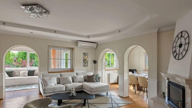 Un salon spacieux avec un canapé d’angle, une table basse et une cheminée. De grandes fenêtres et une porte en arc donnent accès à une autre pièce.