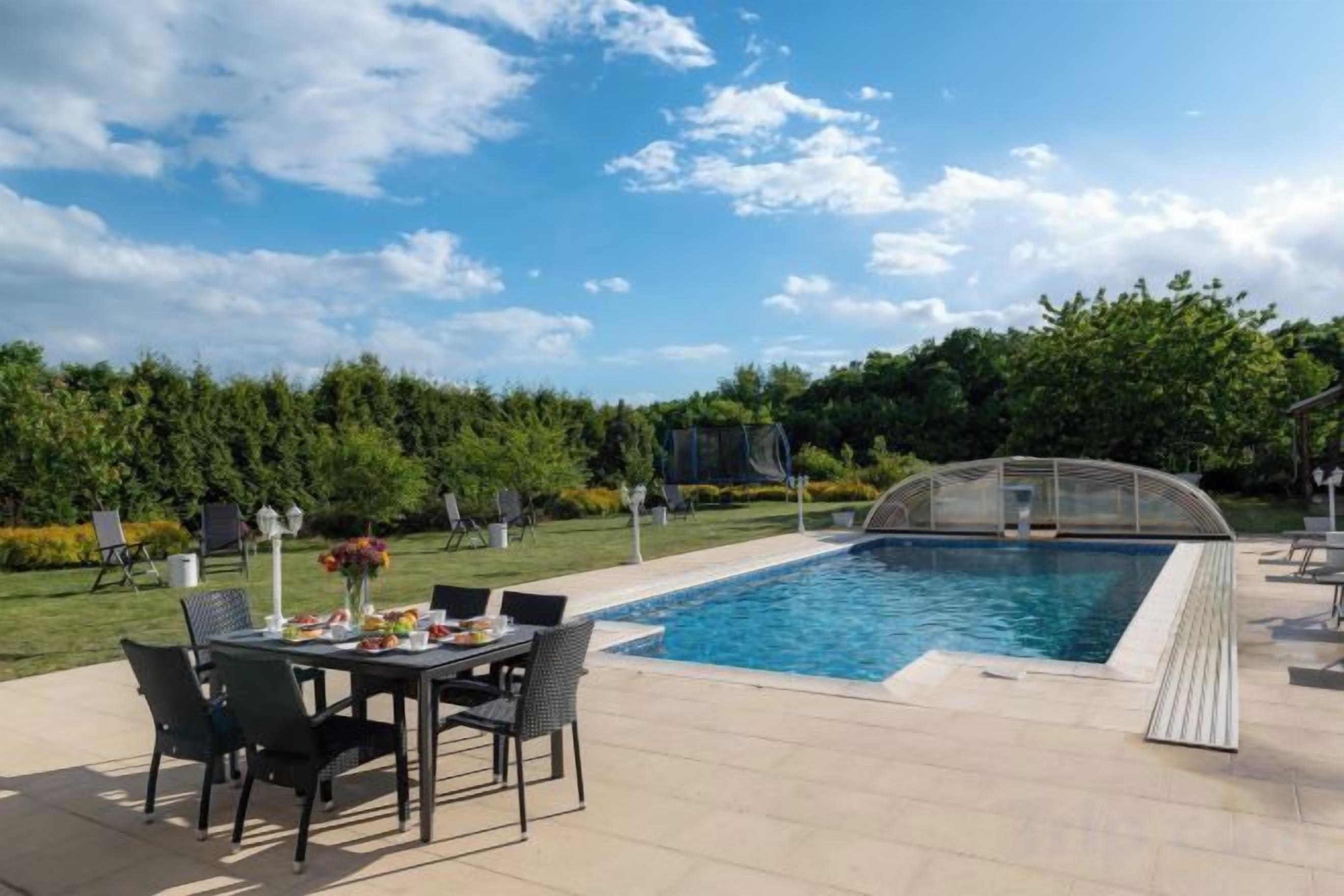 An outdoor swimming pool with a table and chairs, surrounded by a well-maintained garden and trees.