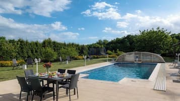 An outdoor swimming pool with a table and chairs, surrounded by a well-maintained garden and trees.