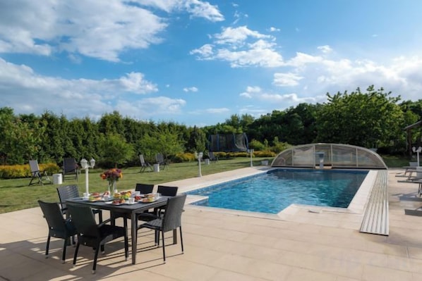 Une piscine extérieure avec une table et des chaises, entourée d’un jardin bien entretenu et d’arbres.