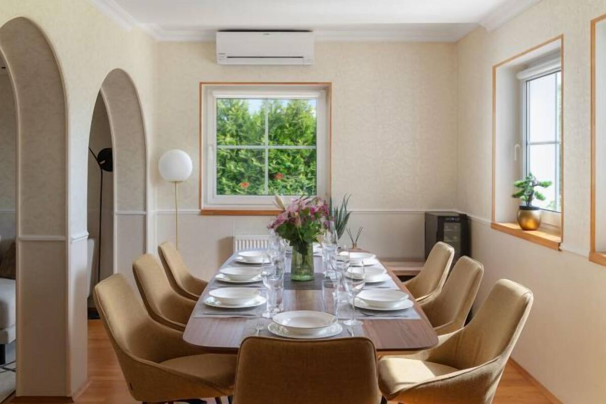 Comedor con una larga mesa dispuesta para seis personas, una ventana con vistas a zonas verdes y un moderno equipo de aire acondicionado en la pared.