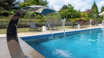 Una piscina con un moderno elemento acuático, rodeada de un jardín ajardinado y mobiliario exterior.