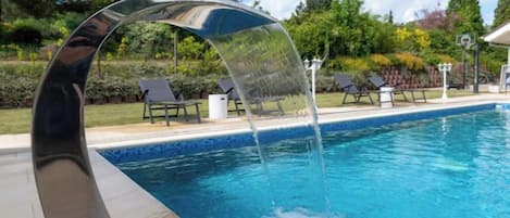 Una piscina con un moderno elemento acuático, rodeada de un jardín ajardinado y mobiliario exterior.