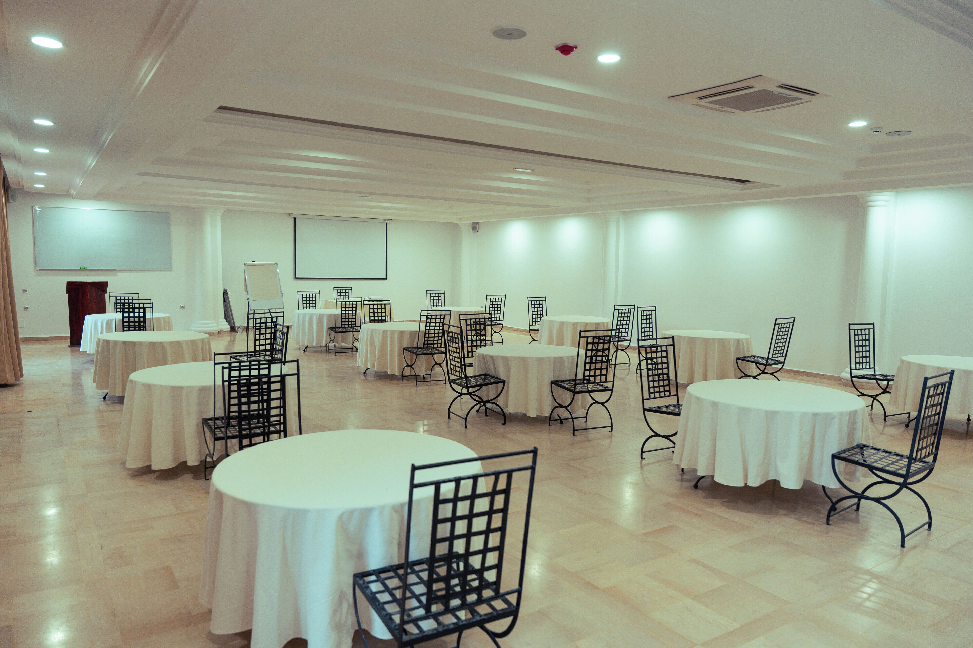 Una sala de conferències amb diverses taules rodones i cadires, parets blanques i una pantalla projectora.