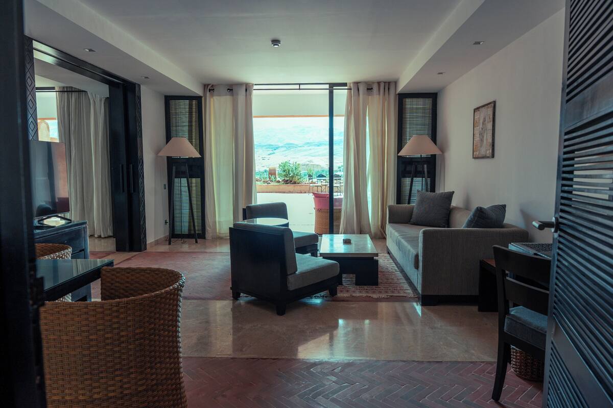 Un salon moderne comprenant un canapé, des fauteuils, une table basse et une table à manger. On peut admirer, depuis le salon, un balcon aménagé avec du mobilier d’extérieur.