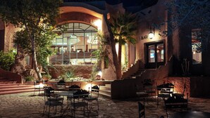 Un patio al aire libre con piso de piedra, mesas y sillas, rodeado de árboles y un edificio con un amplio ventanal y un letrero de bar.