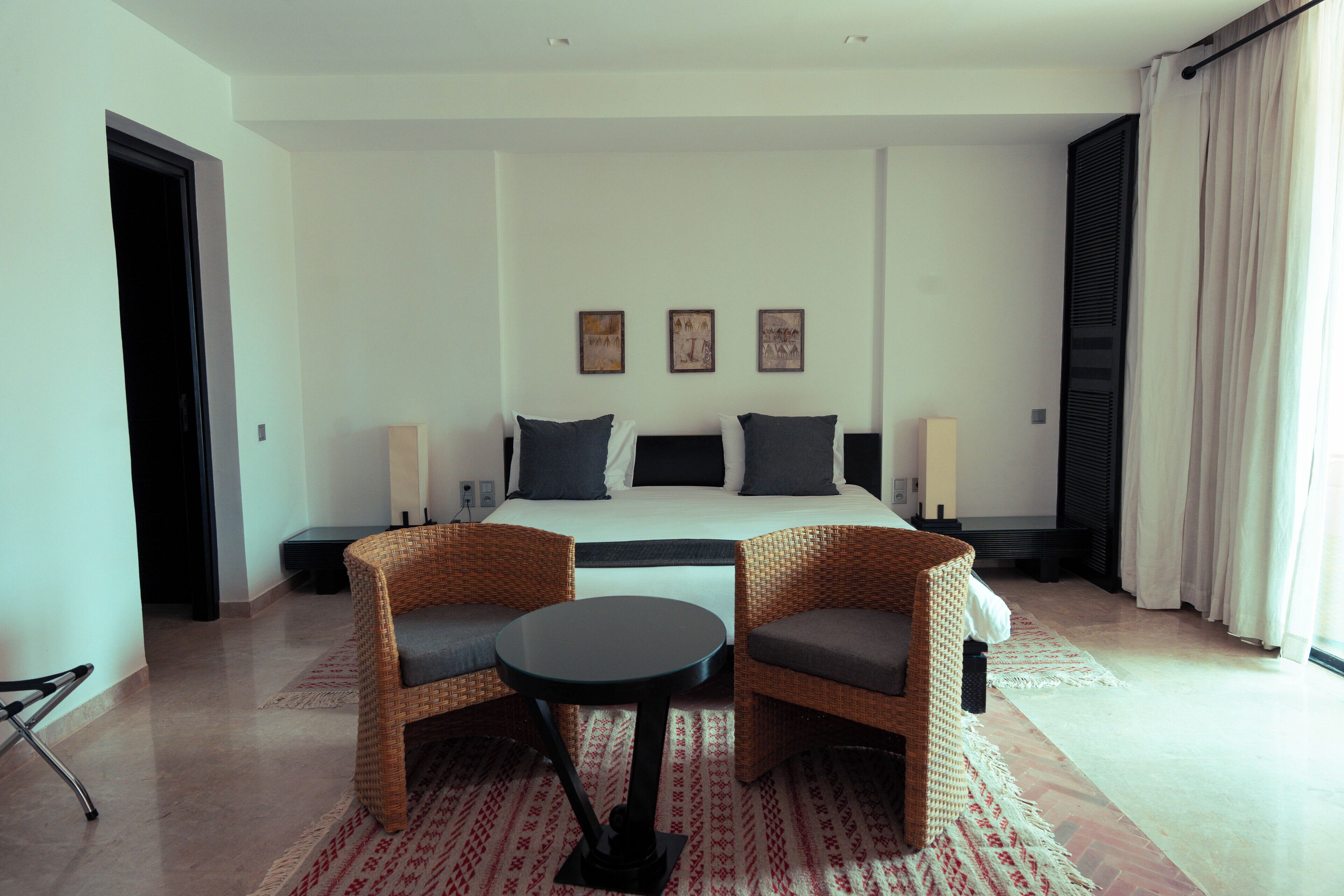 Une chambre d’hôtel avec un lit, deux chaises, une petite table et une fenêtre avec des rideaux.