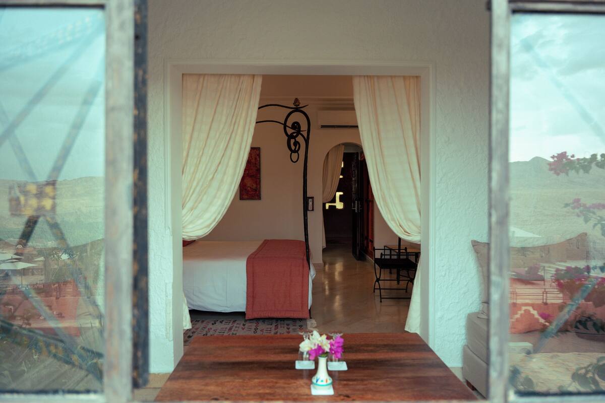 Une chambre à coucher avec un lit, une table de chevet ornée d’un vase, et une vue par la fenêtre.