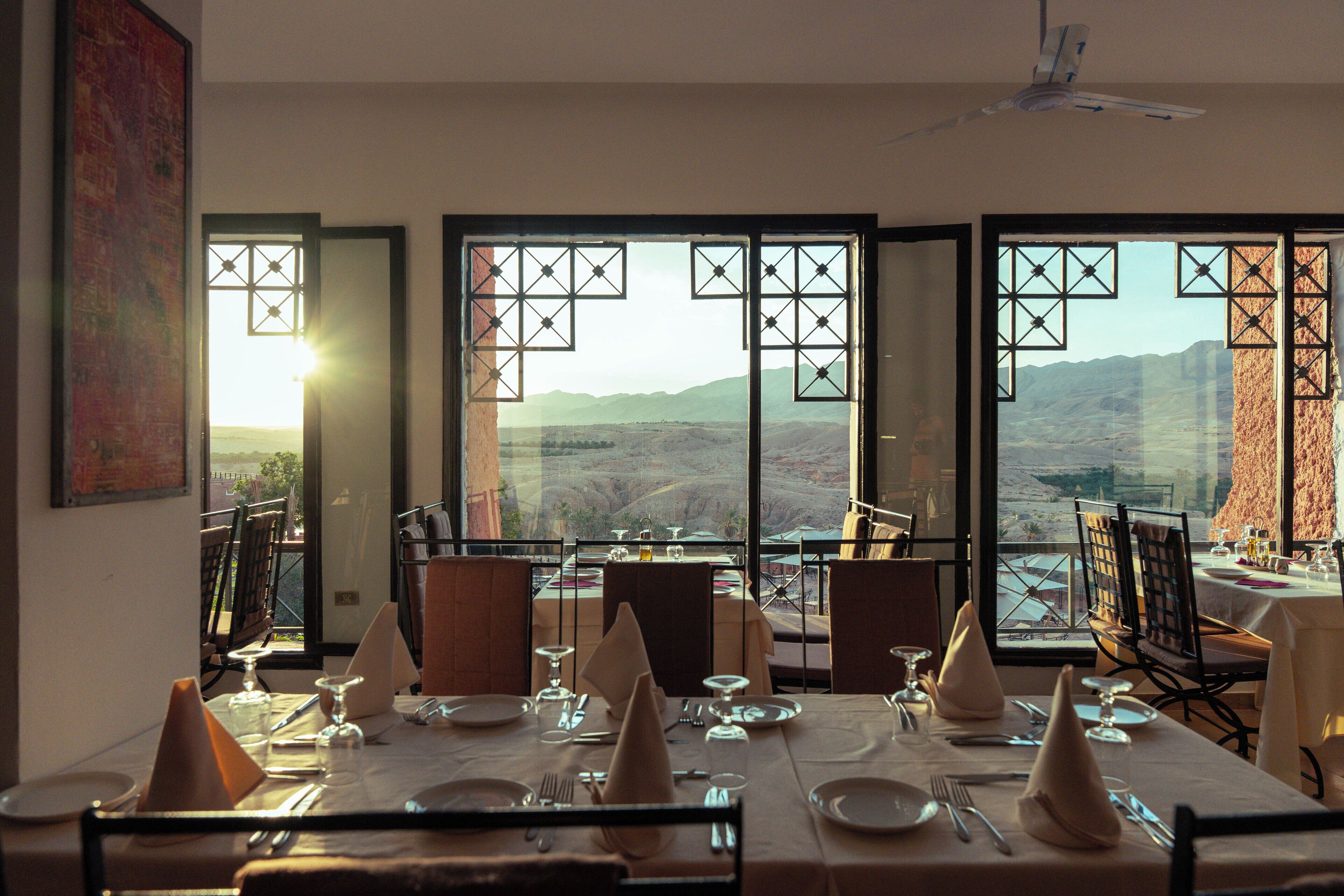 A dining area with tables set for two, large windows with geometric patterns, and a view of a mountainous landscape.