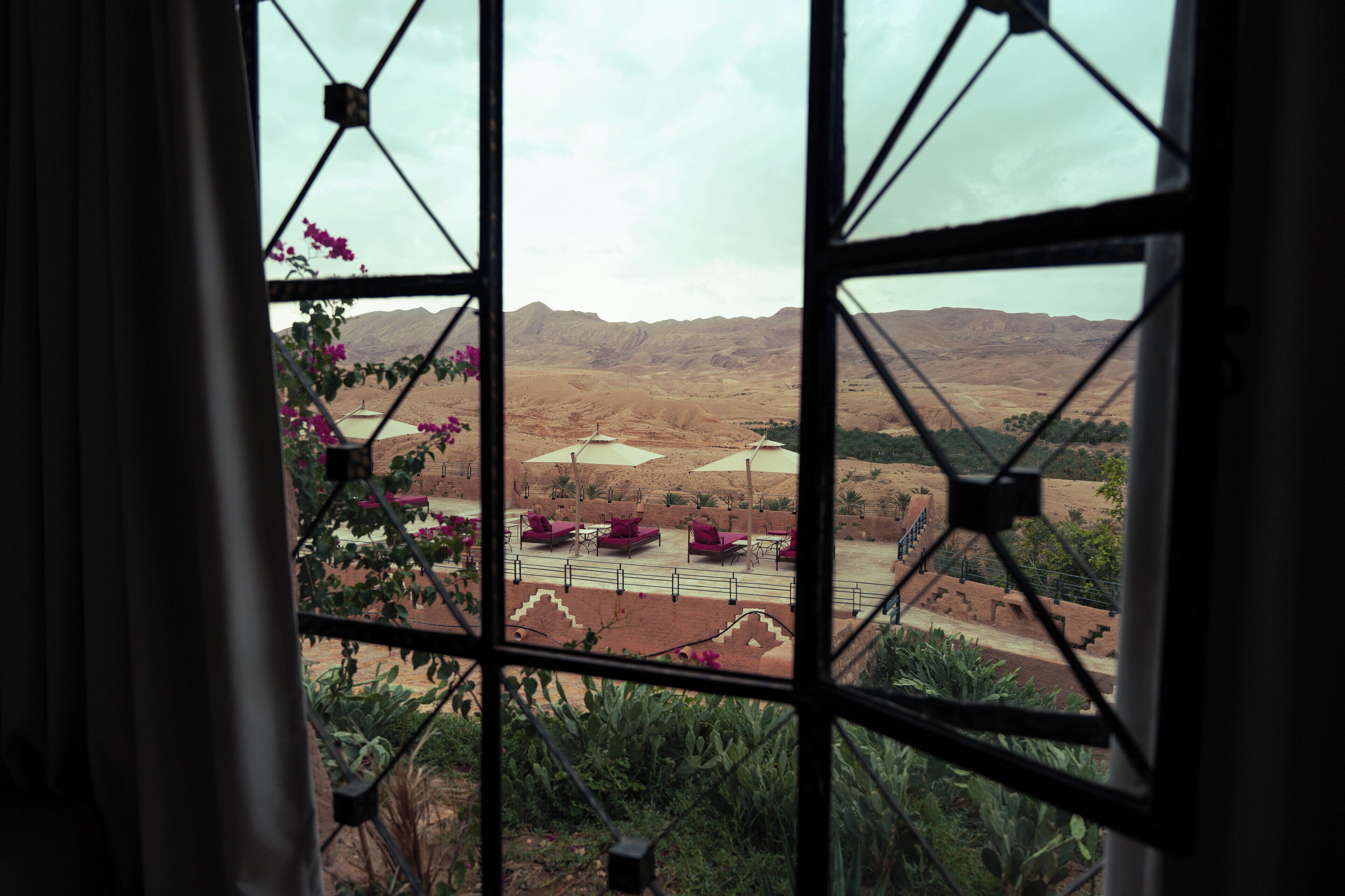 A view through a window with a geometric pattern, featuring a desert landscape with distant mountains, a paved area with umbrellas, and some greenery.