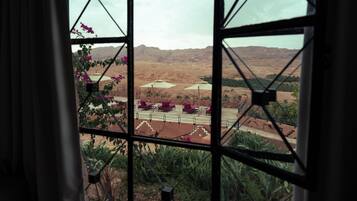 A view through a window with a geometric pattern, featuring a desert landscape with distant mountains, a paved area with umbrellas, and some greenery.