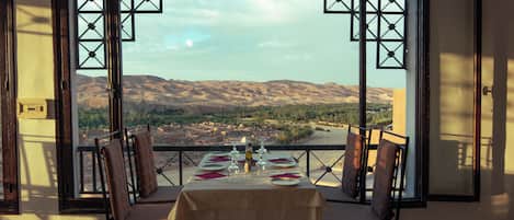 Comedor con una mesa dispuesta para cuatro personas, con vistas a un paisaje montañoso a través de puertas abiertas.