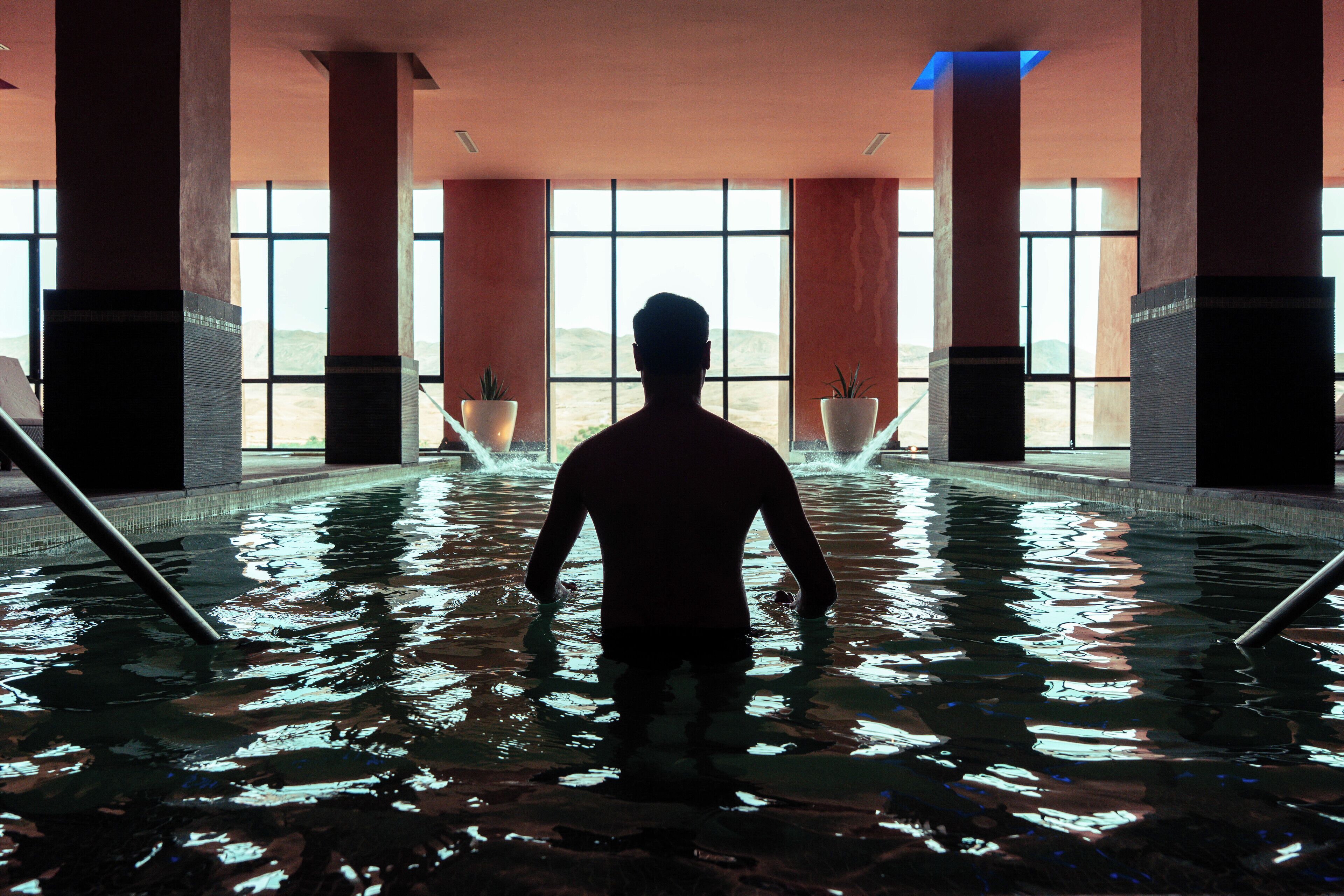 A person standing in a large indoor pool with a view of the outdoors through large windows.