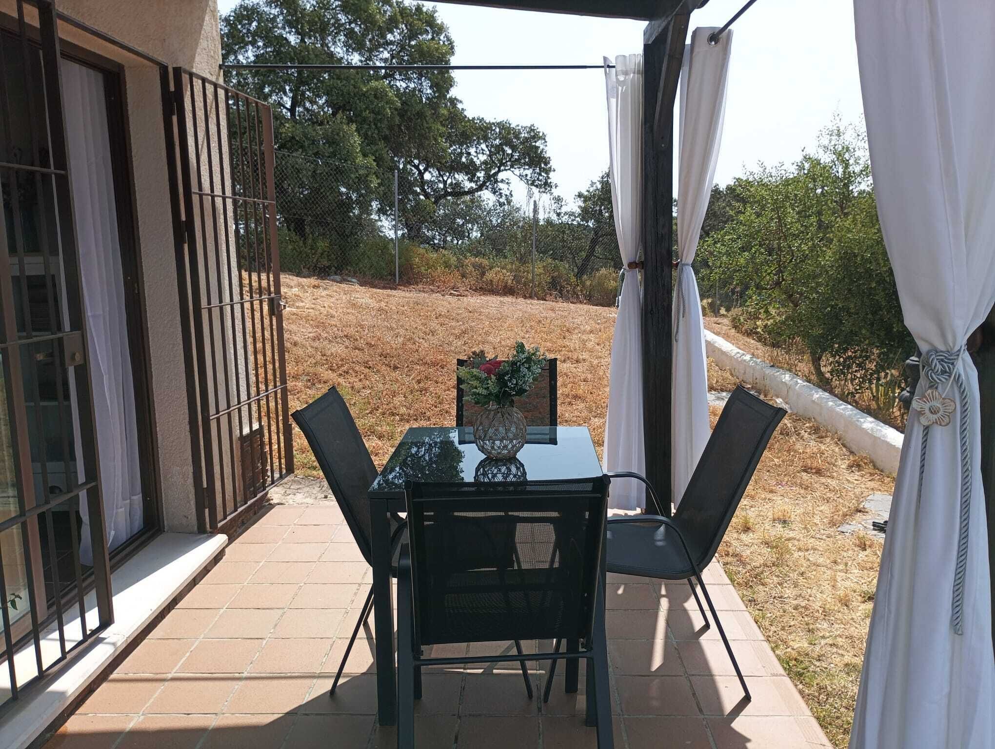 An outdoor patio with a table and chairs, a glass door, and a view of a grassy area with trees.