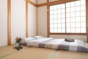 A traditional Japanese room with tatami flooring, a low bed, a window with shoji screens, and a small vase with flowers. - Younoyado (Osaka)