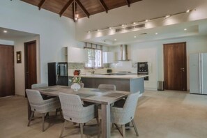 Een moderne keuken met een centraal eiland, een eetgedeelte met tafel en stoelen, en een hoge plafond met zichtbare balken.