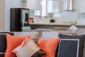 Ein Wohnzimmer mit einer Couch und Kissen, im Hintergrund eine Küchenzeile und ein Esstisch.