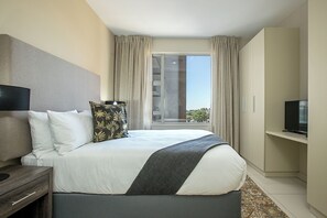 Ein Hotelzimmer mit einem großen Bett, einem Fernseher, einem Schreibtisch und einem Fenster mit Vorhängen.