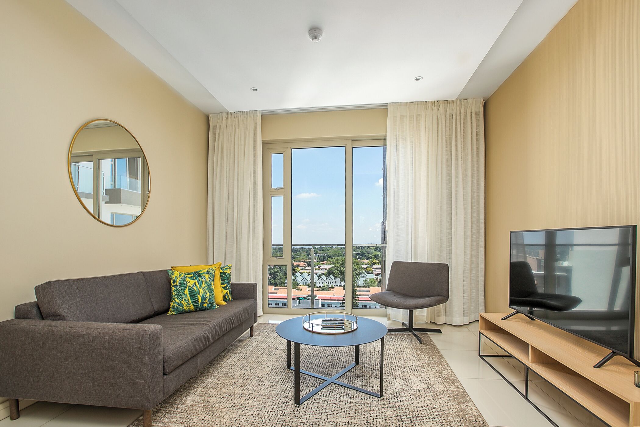 Ein modernes Wohnzimmer mit Sofa, Sessel, Couchtisch, Fernseher und großen Fenstern mit Blick auf die Stadt.