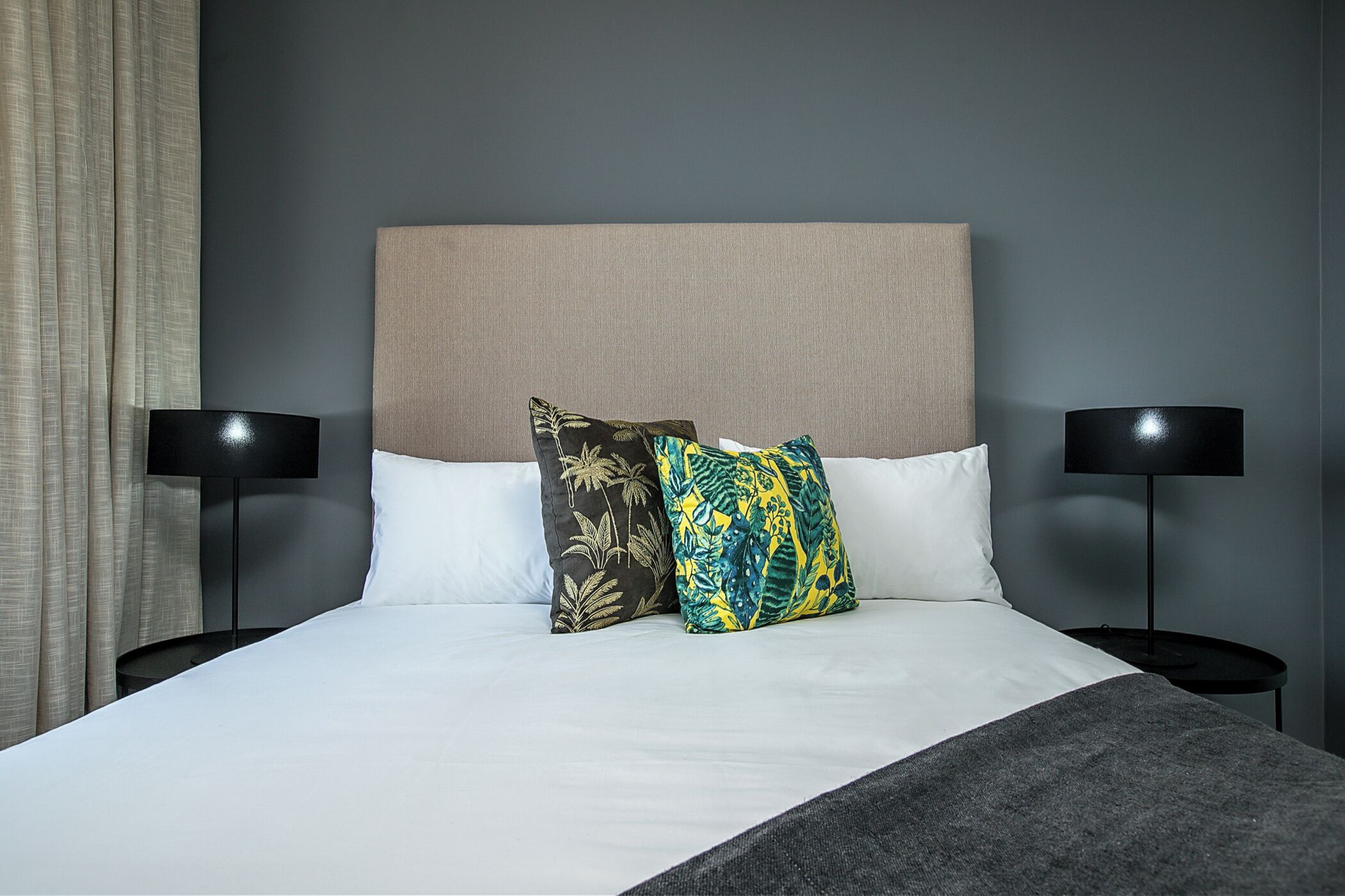 A neatly made bed with two pillows, a grey blanket, and two black bedside tables with lamps.