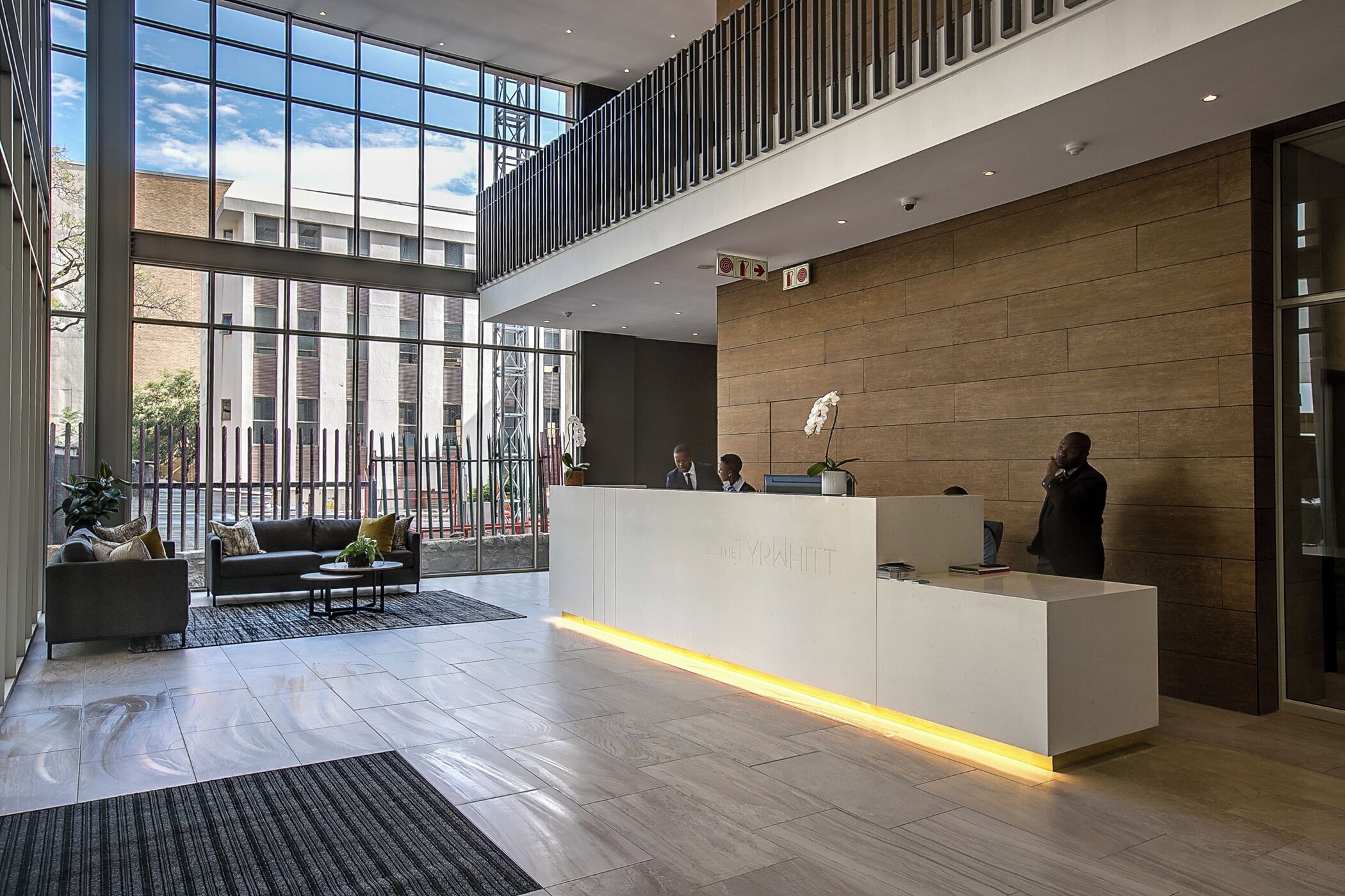 A modern reception area with a white reception desk, a sofa, and a coffee table. There are large windows providing natural light and a view of the outside.