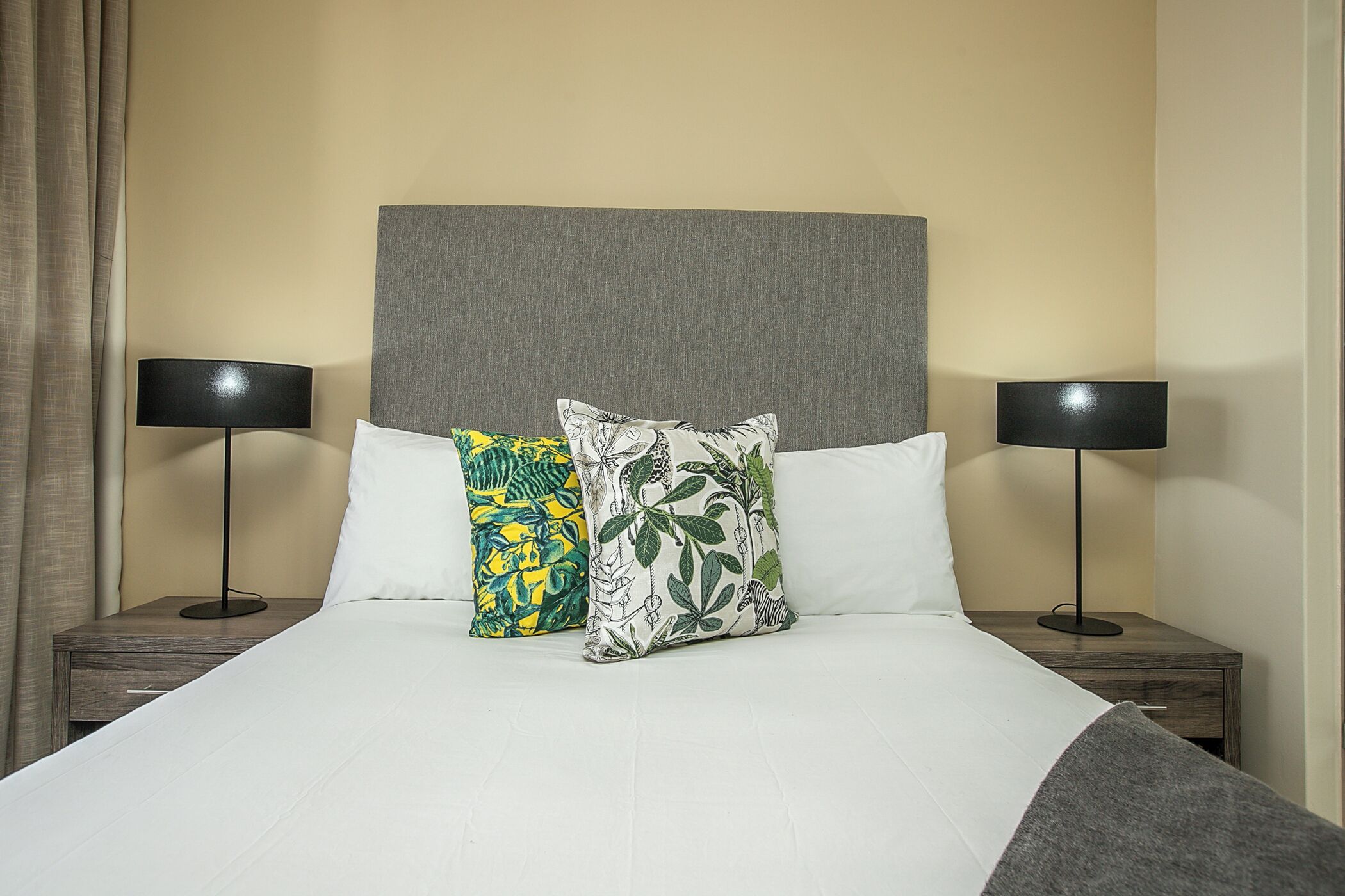 Una cama bien hecha con cabecera gris, dos lámparas de noche y dos almohadones decorativos.