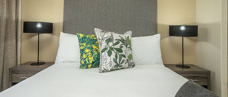 Una cama bien hecha con cabecera gris, dos lámparas de noche y dos almohadones decorativos.