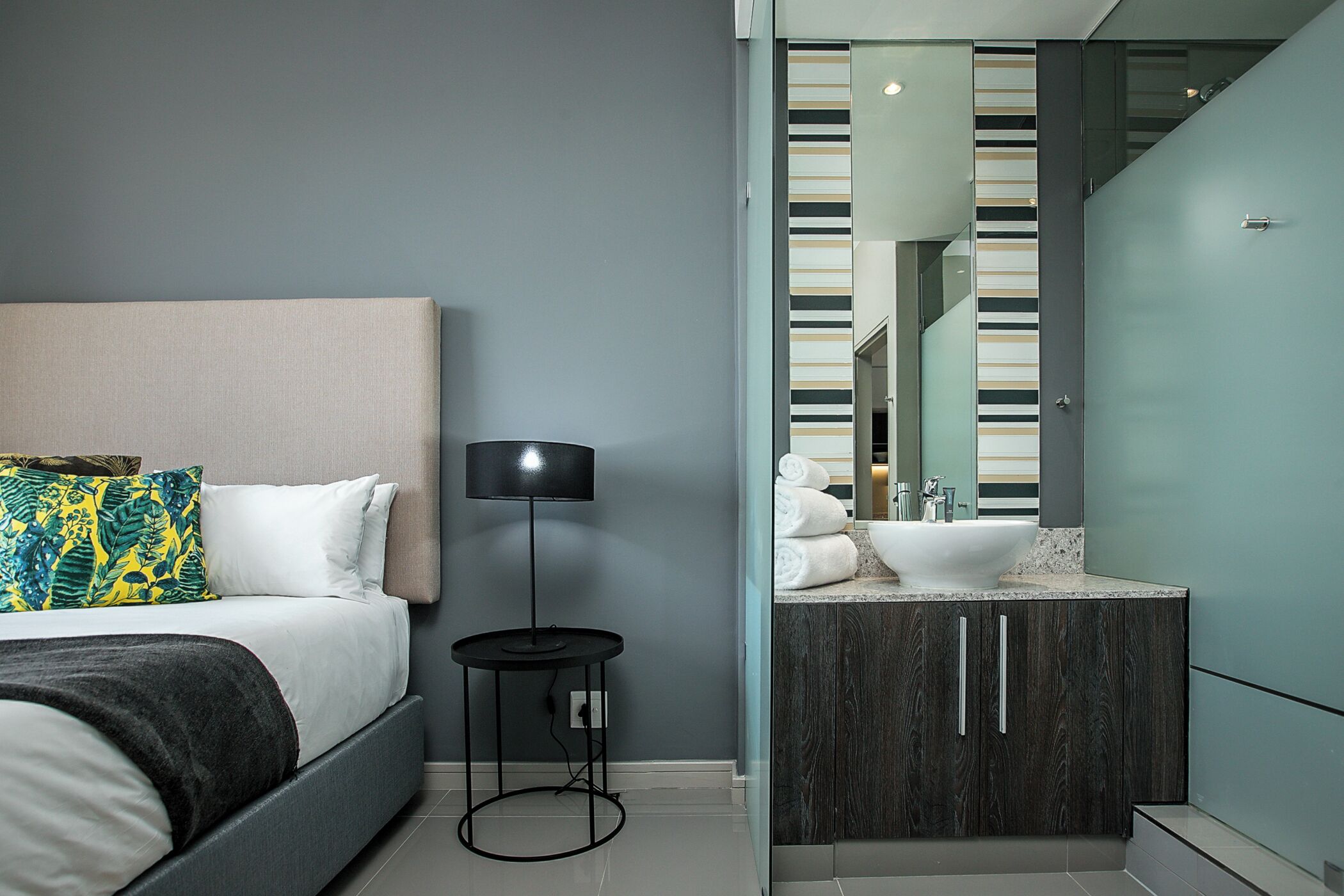 A modern bedroom with a bed, bedside lamp, and a bathroom with a sink and mirror.