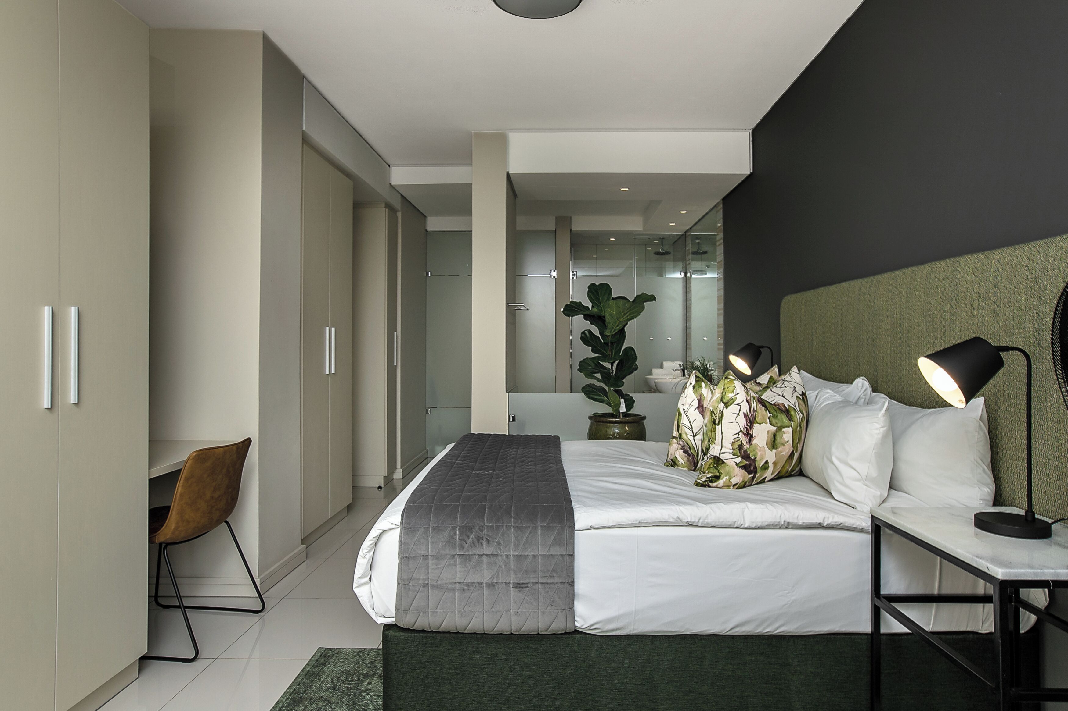 A modern bedroom with a bed, bedside table, lamp, and a large potted plant.