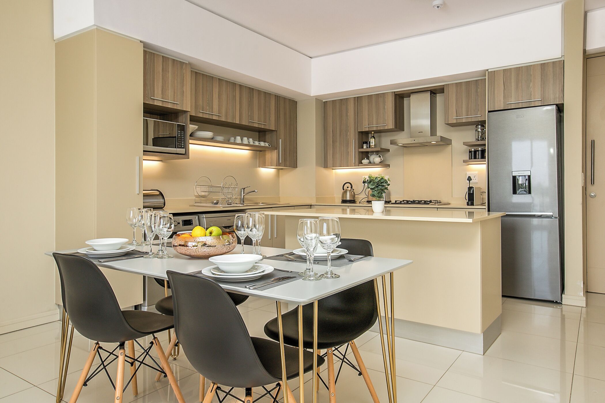A modern kitchen with wooden cabinets, a stainless steel refrigerator, and a dining area with a table set for four.
