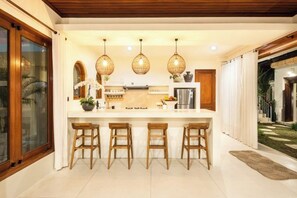 A modern kitchen with a central island, wooden stools, and pendant lighting. - Villa Casa Toya (Bali)
