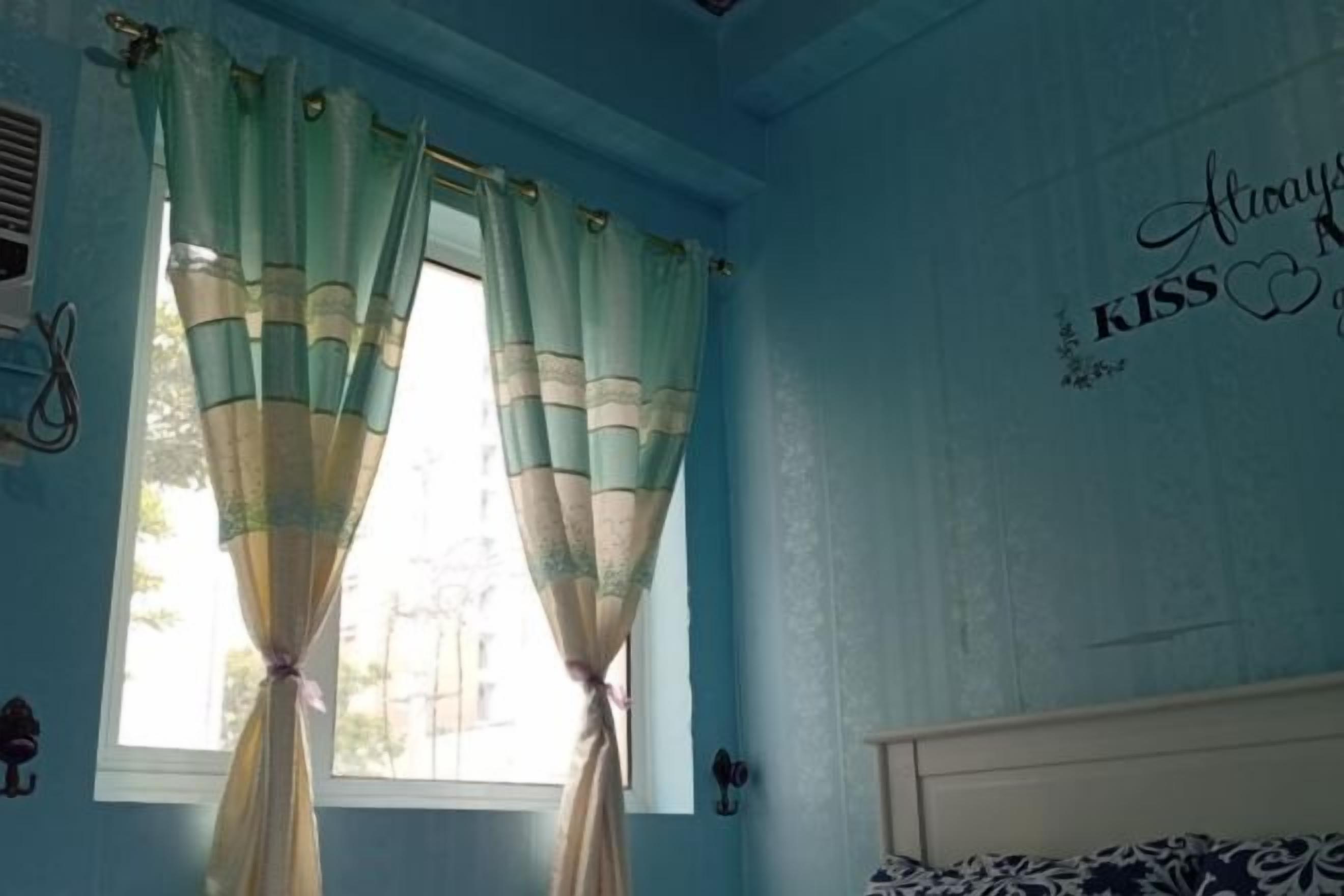 A room with a window, curtains, a bed, and a wall with a decorative phrase.