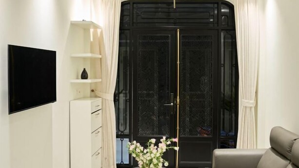 A modern living room with a flat-screen TV, a white cabinet with drawers, a vase with flowers, and a large black door with decorative glass panels. - Home by La Sinfonía Trieu Viet Vuong (Hanoi)