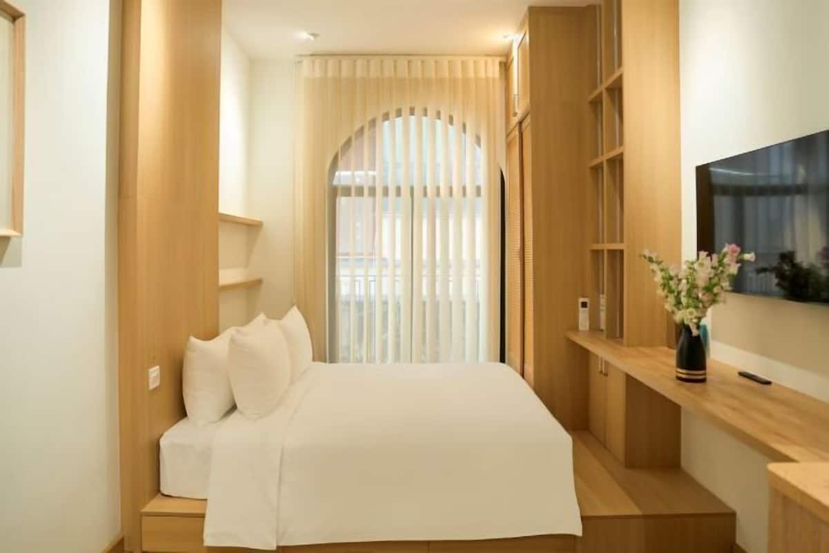 A modern hotel room with a bed, wooden shelving, a TV, and a window with curtains.