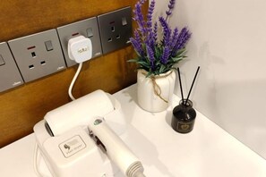 Valkoisella tasolla on hiustenkuivaaja, pistorasia ja maljakko, jossa on laventelinkukkia.