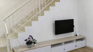 Egy lépcső, fehér korláttal, egy falra szerelt, síkképernyős televízió, egy fából készült, fiókos konzolasztal és két virágtartóban elhelyezett növény.