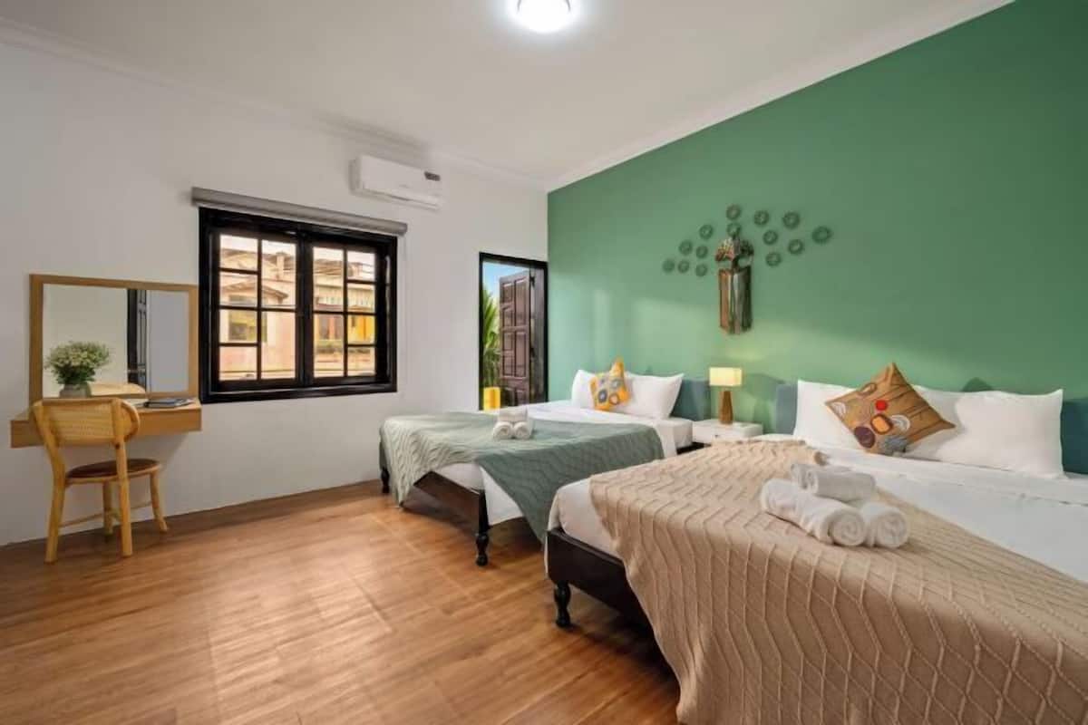 A hotel room with two beds, a desk with a chair, a window, and a green wall with decorative elements.